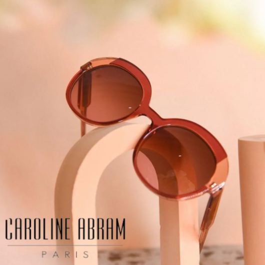 Red round sunglasses on a curved beige pedestal against a soft peach background, with "CAROLINE ABRAM PARIS" text.