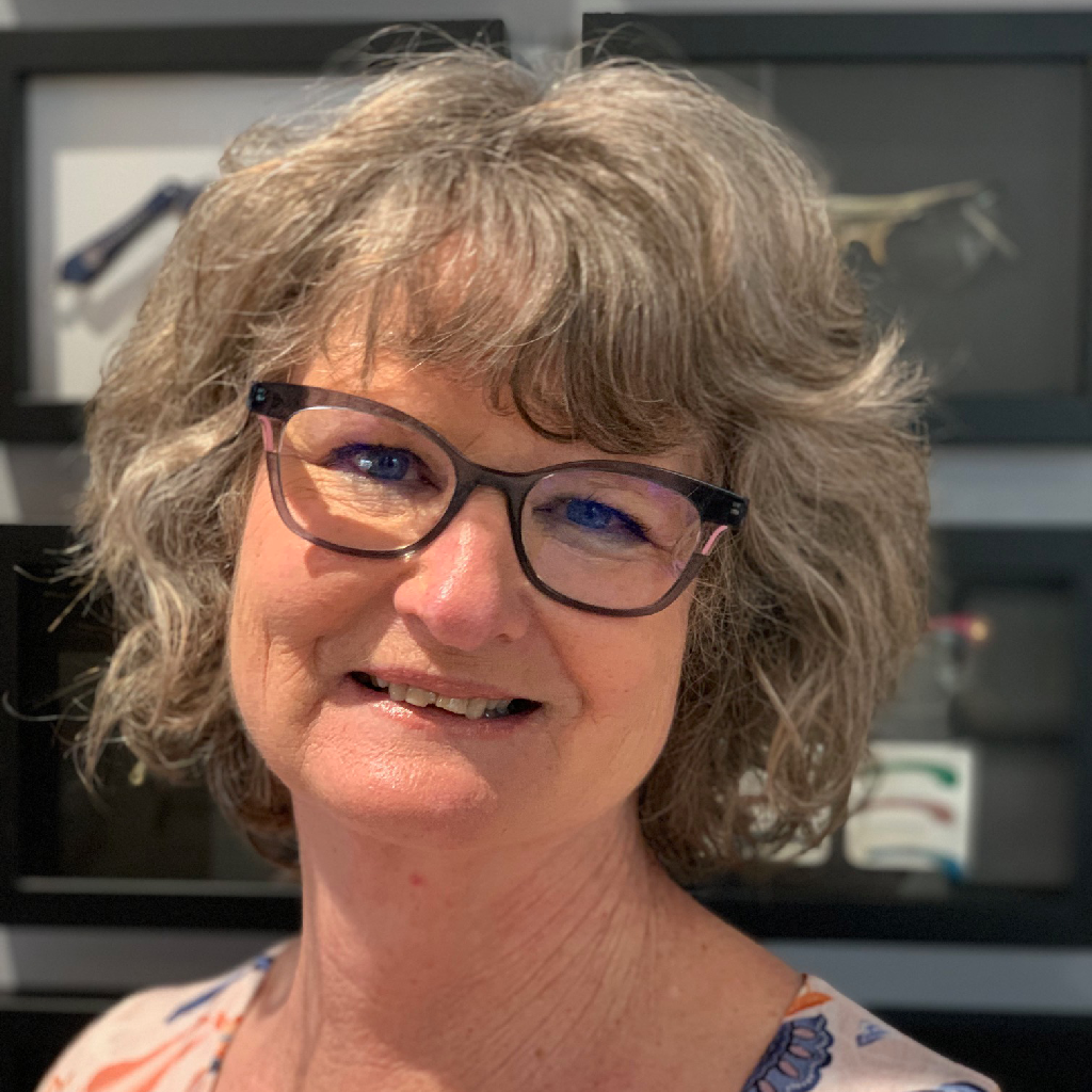 Smiling person with gray hair and glasses in front of shelves and framed artwork