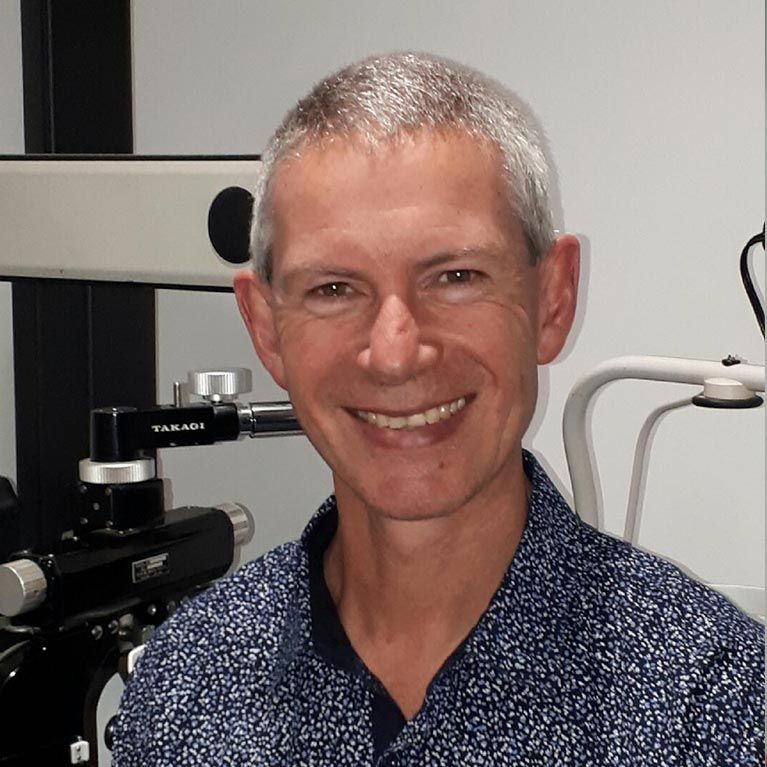 Smiling man in a blue patterned shirt standing beside a microscope in a lab or clinic.