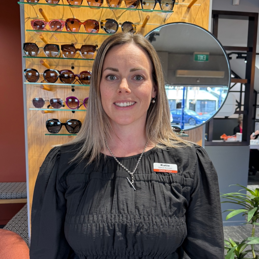 Person in a black blouse stands in an eyewear store, with sunglasses displayed behind her.
