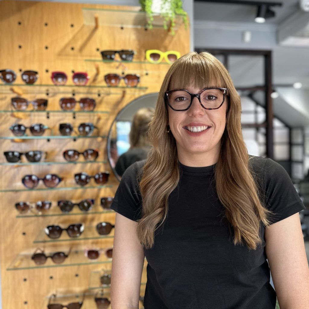 Woman smiling in glasses inside an eyeglasses shop, with frames displayed on the wall.