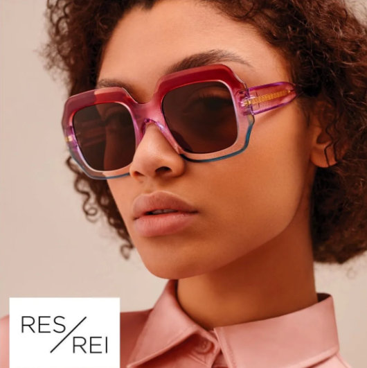 Fashion model wearing oversized pink sunglasses and a blush satin shirt on a neutral background.