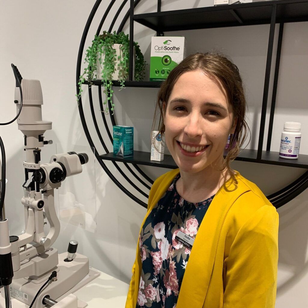 Smiling woman in a yellow jacket stands beside a microscope in an optometry office.