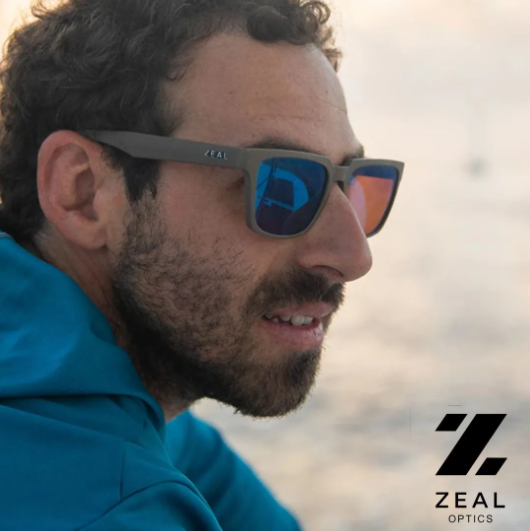 Man in blue jacket wearing reflective sunglasses near water at sunset, with ZEAL logo in the corner