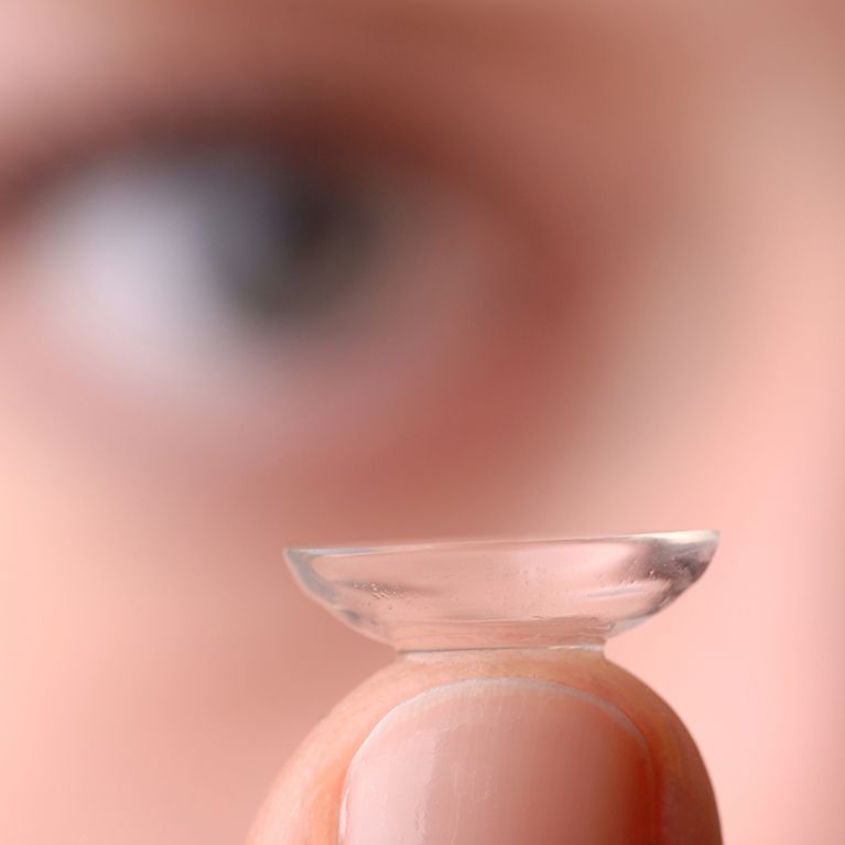 A contact lens held on a fingertip, with a blurred eye in the background.