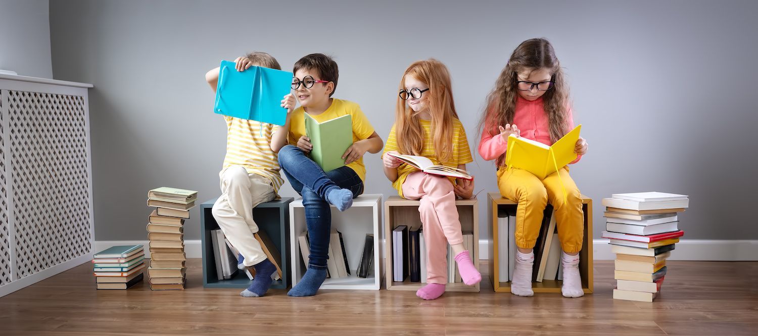 Four children sit on stools reading colorful books in a bright classroom-like room