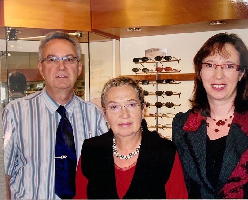 Three people standing indoors, wearing business attire in front of a display of eyeglasses.