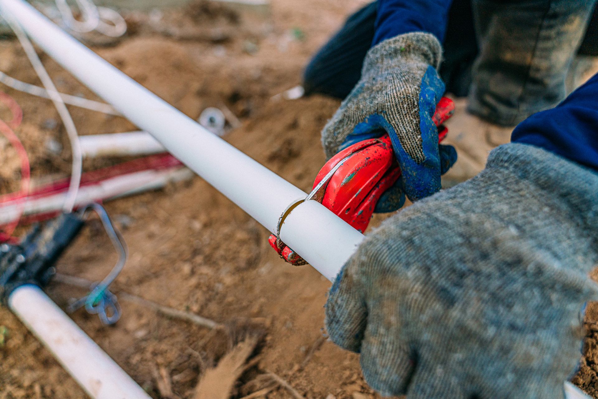 Hands wearing gloves cutting white PVC pipe with a red cutter, outdoors.