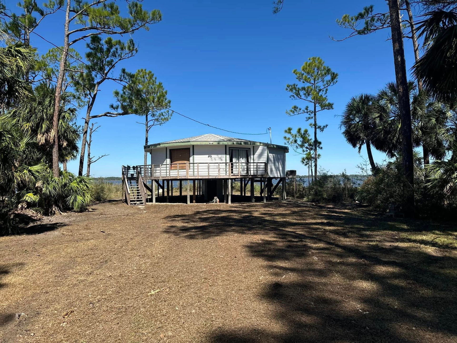 Small white stilt house on a sandy lot with trees and a bright blue sky