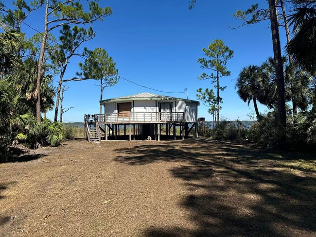 Small white cabin on stilts in a sunny clearing, surrounded by sparse trees under a blue sky.