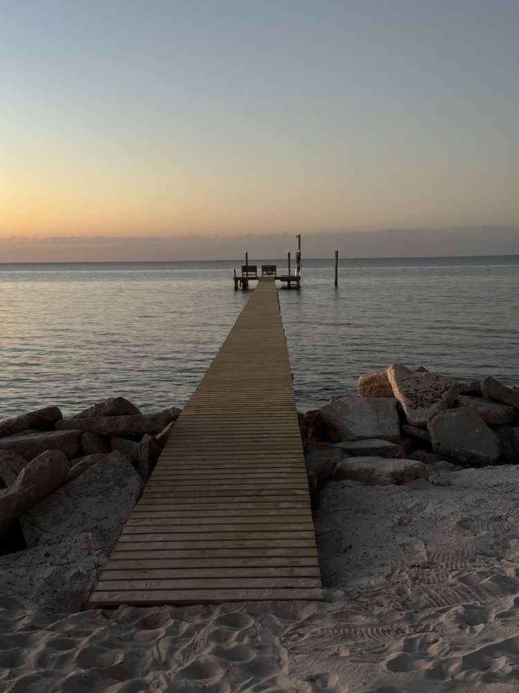 Wooden pier leading to a calm sea at sunset, with rocks and a soft orange horizon.