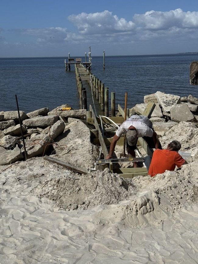 Two workers on a sandy shore repairing a wooden pier extending into calm blue water