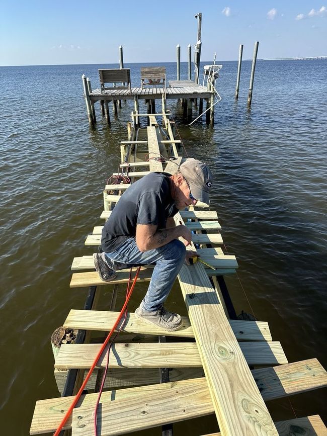 Man working on a wooden dock over calm water, with a larger pier in the background.