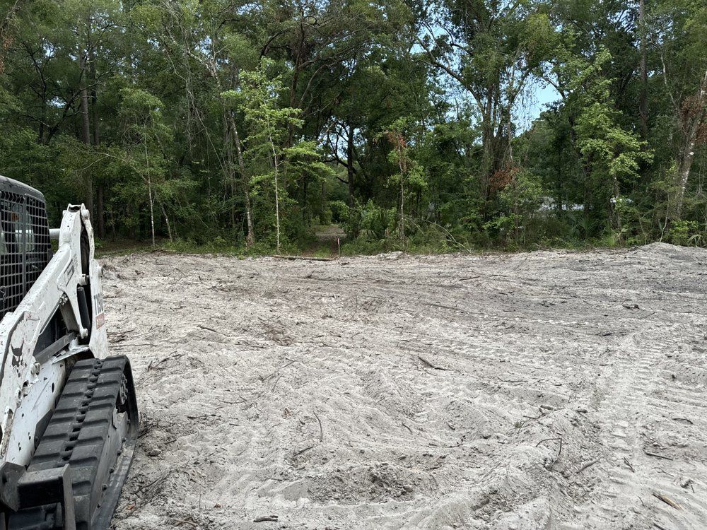Construction site with a white excavator beside a cleared dirt lot and trees in the background