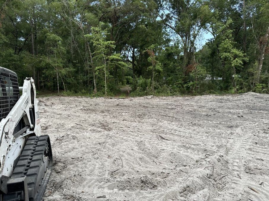 Construction site with a tracked skid steer on a cleared dirt lot beside trees
