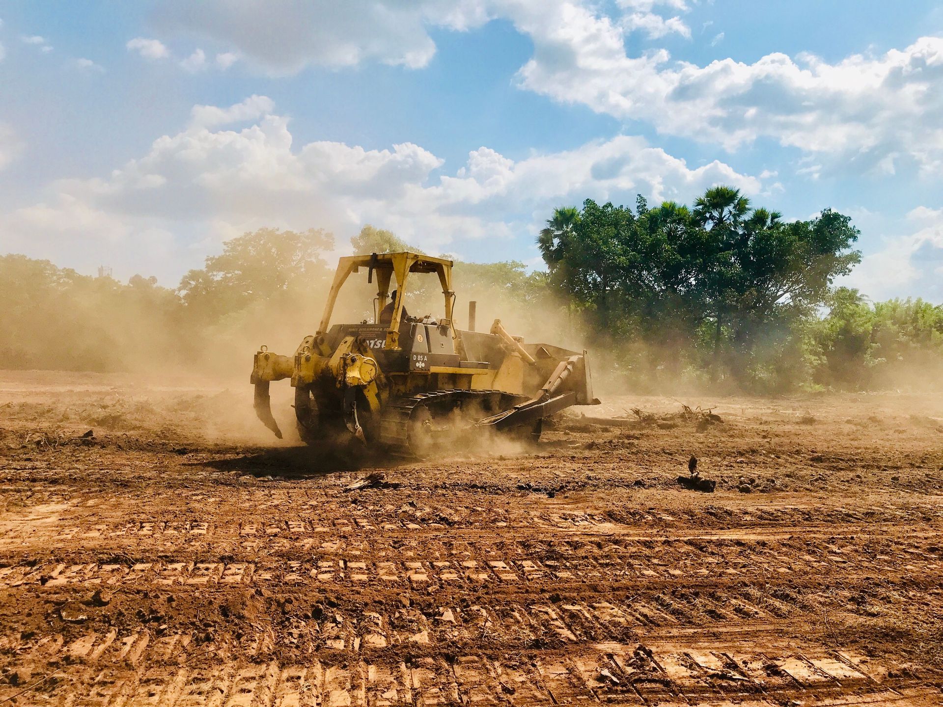 Yellow tractor leveling a dusty dirt field under a blue sky with trees in the background