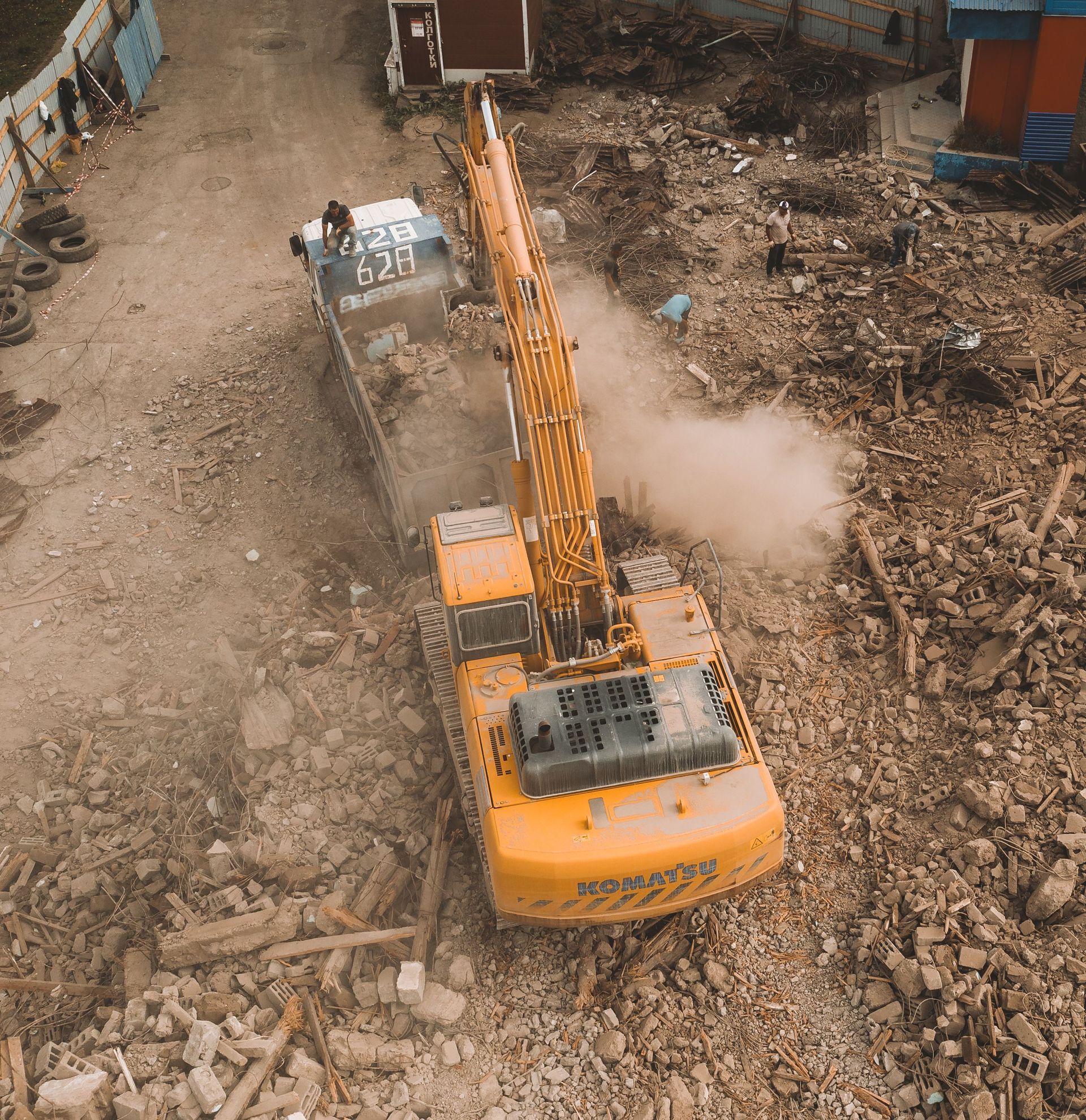 Orange excavator clearing rubble in a demolition site, with dust rising around it.