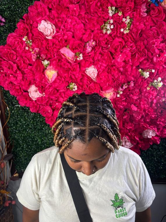 A person with braided hair styled in square parts, featuring blonde tips, against a vibrant red rose backdrop.