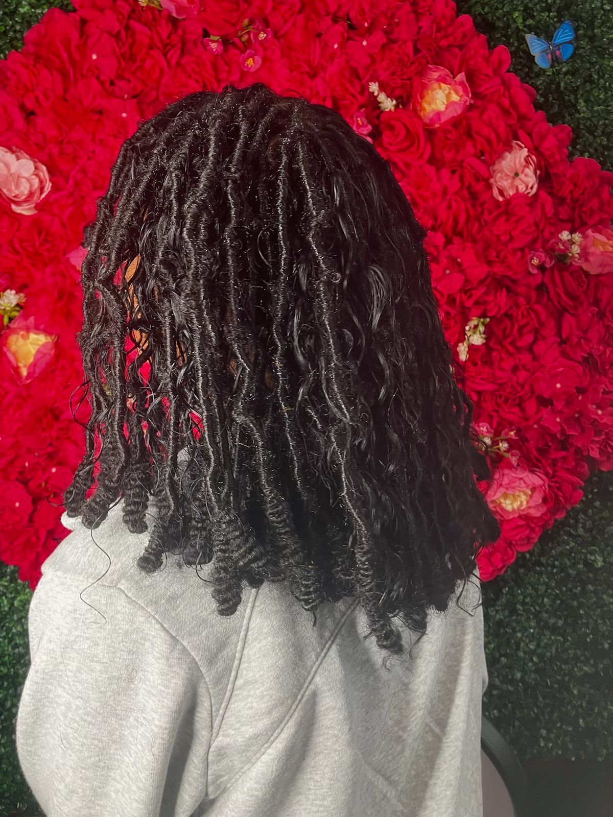 Back view of a person with shoulder-length, dark, coiled locs in front of a wall of red flowers.