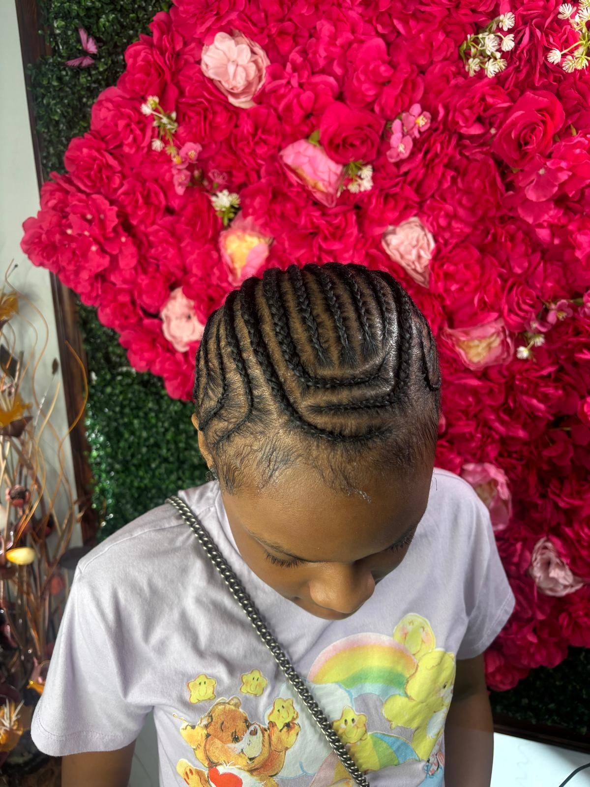 A person with intricate braided cornrows arranged in a curved pattern, standing in front of a wall of red flowers.