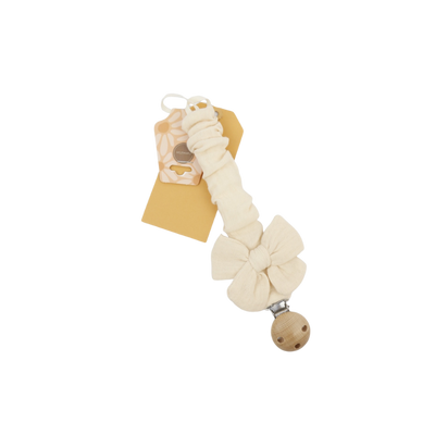 Een crèmekleurige stoffen speenclip met een strikje en een houten bevestiging, gepresenteerd met een papieren productlabel.