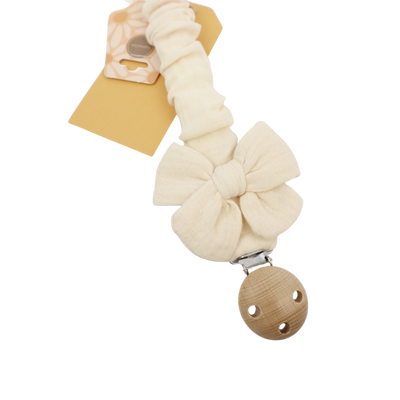 Een crèmekleurige stoffen speenclip met een strikje en een houten bevestiging, gepresenteerd met een papieren productlabel.