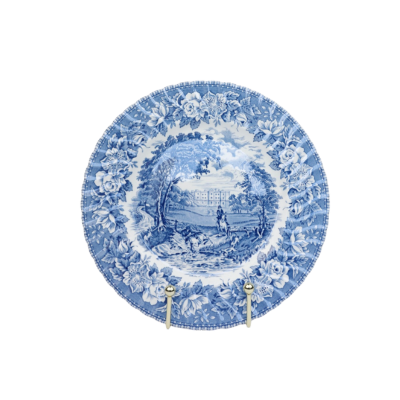 Een blauw-wit decoratief keramisch bord met een landelijk landschap, gepresenteerd op een klein zilveren voetstuk.