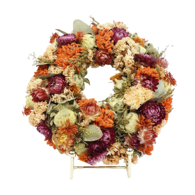 Een ronde krans van gedroogde oranje, paarse en crèmekleurige bloemen rust op een klein gouden standaardje.