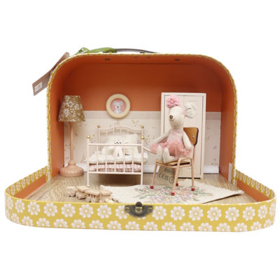 Een muizenpop in een tutu op een stoel in een koffer die is ingericht als een miniatuurslaapkamer met een bed en een lamp.