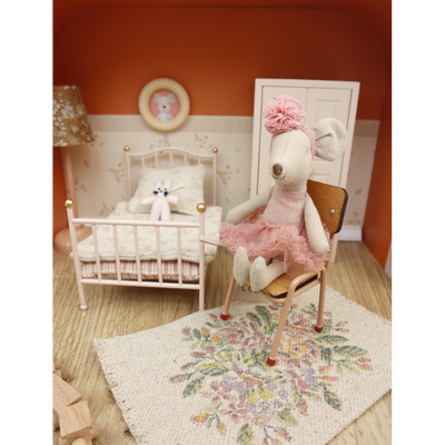 Een muizenpop in een tutu op een stoel in een koffer die is ingericht als een miniatuurslaapkamer met een bed en een lamp.