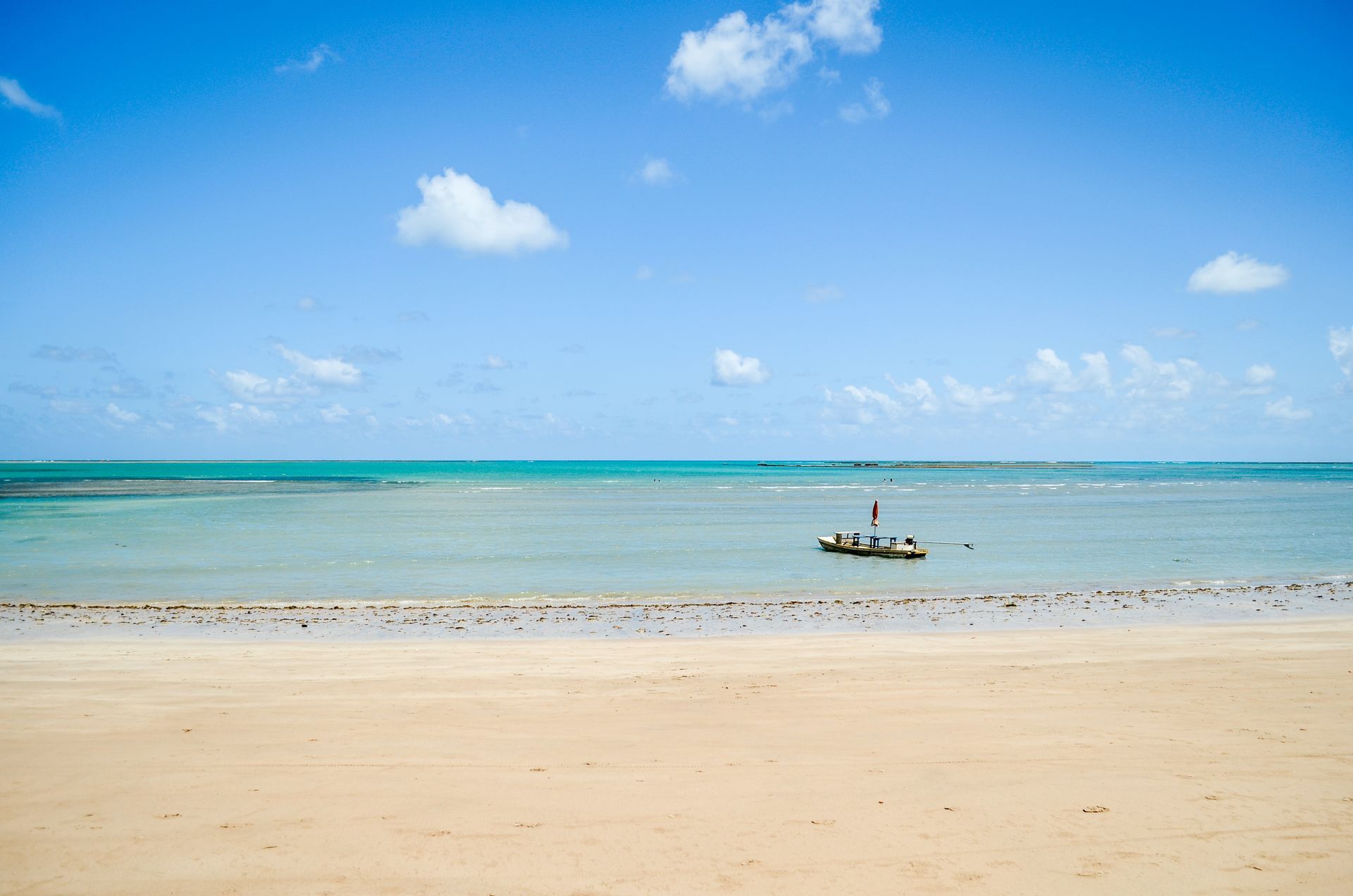 A areia da praia encontra o oceano turquesa sob um céu azul brilhante com nuvens fofas. Um barco está ao longe.