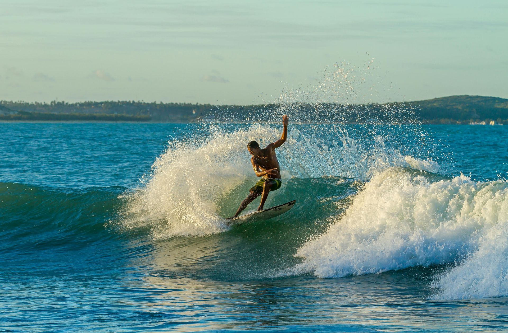 Surfista deslizando sobre uma onda, braços erguidos, rodeado por respingos, contra um mar azul e a costa.