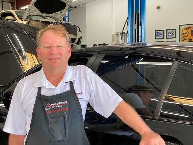 Mechanic standing beside a black car in an auto repair shop, wearing a white shirt and dark apron.