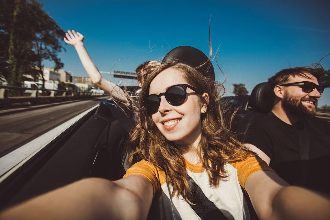 Two people smiling in a convertible on a sunny drive, one waving out the window.