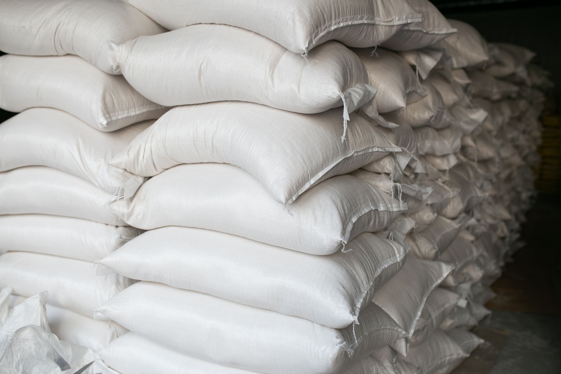 Pile of white sacks, possibly filled with flour, stacked in a warehouse or storage area.