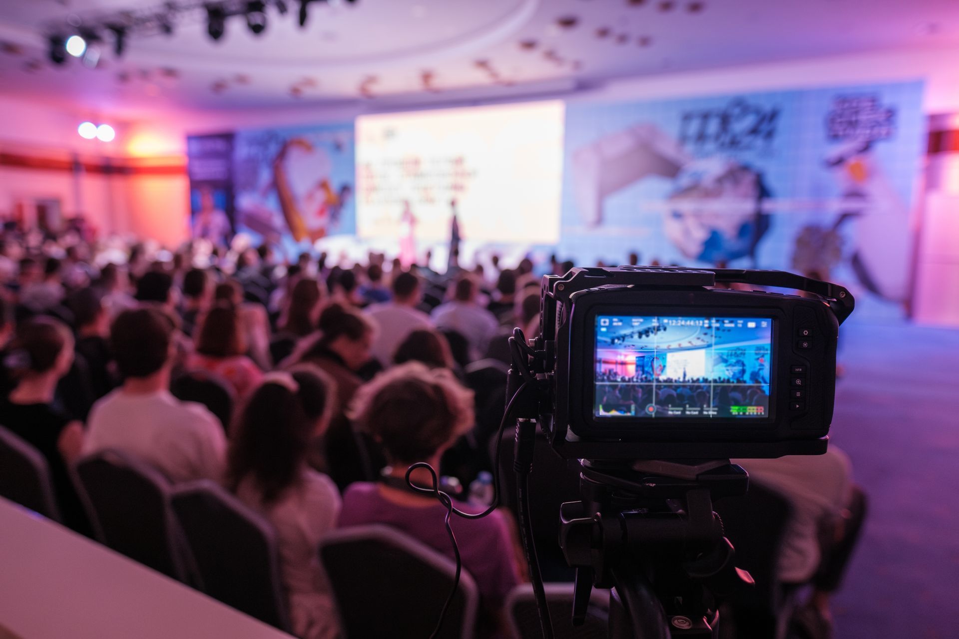 Camera recording a large audience in a brightly lit conference hall with a stage and presentation screen.