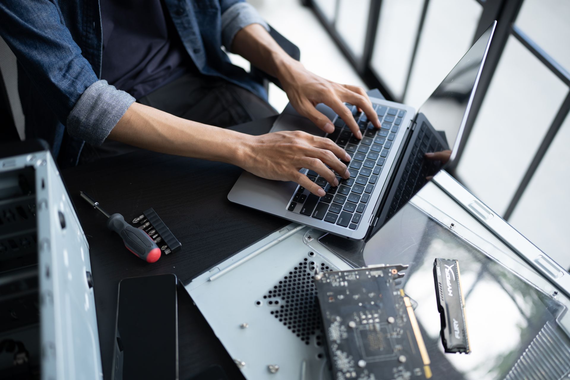 Hands typing on a laptop at a desk with computer parts and tools nearby