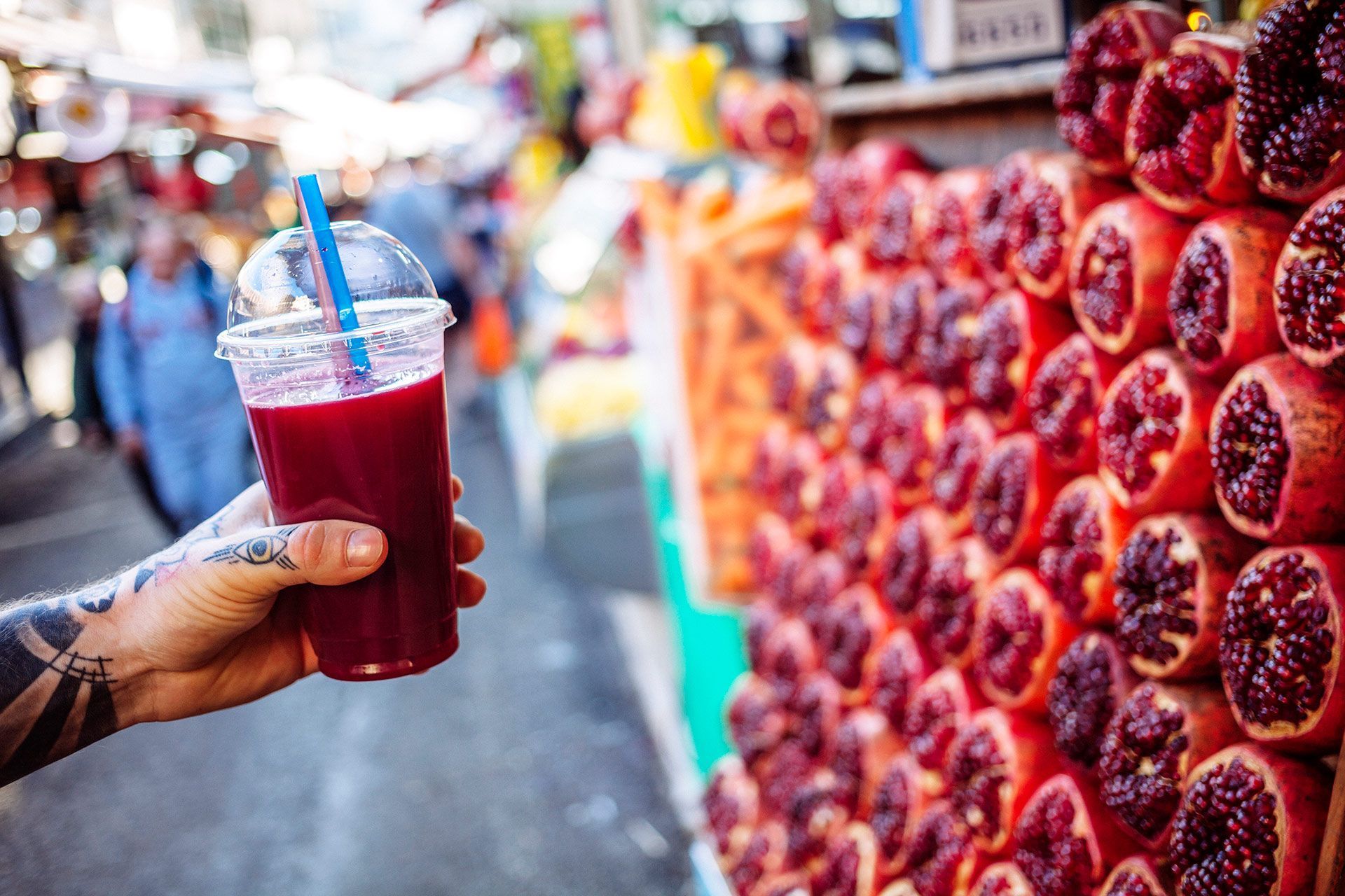 Hand holding a red berry drink with a straw beside a market stall stacked with fresh berries