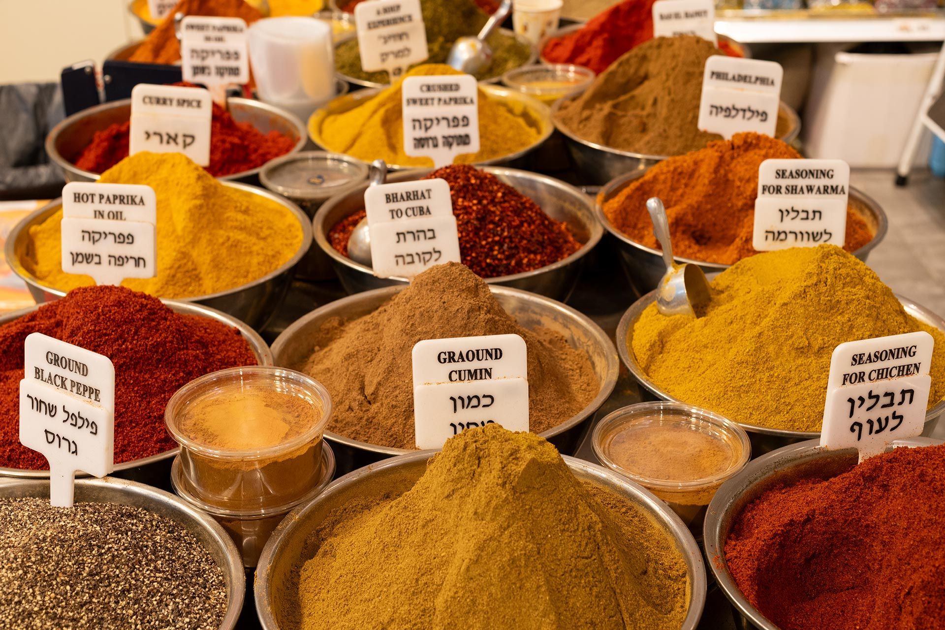 Assorted colorful spices in bowls at a market, with small labeled signs in Hebrew.