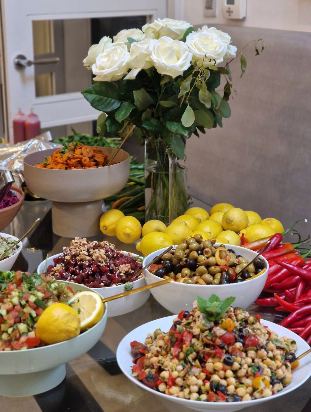 Buffet table with bowls of colorful salads, lemons, red peppers, and white roses in a vase