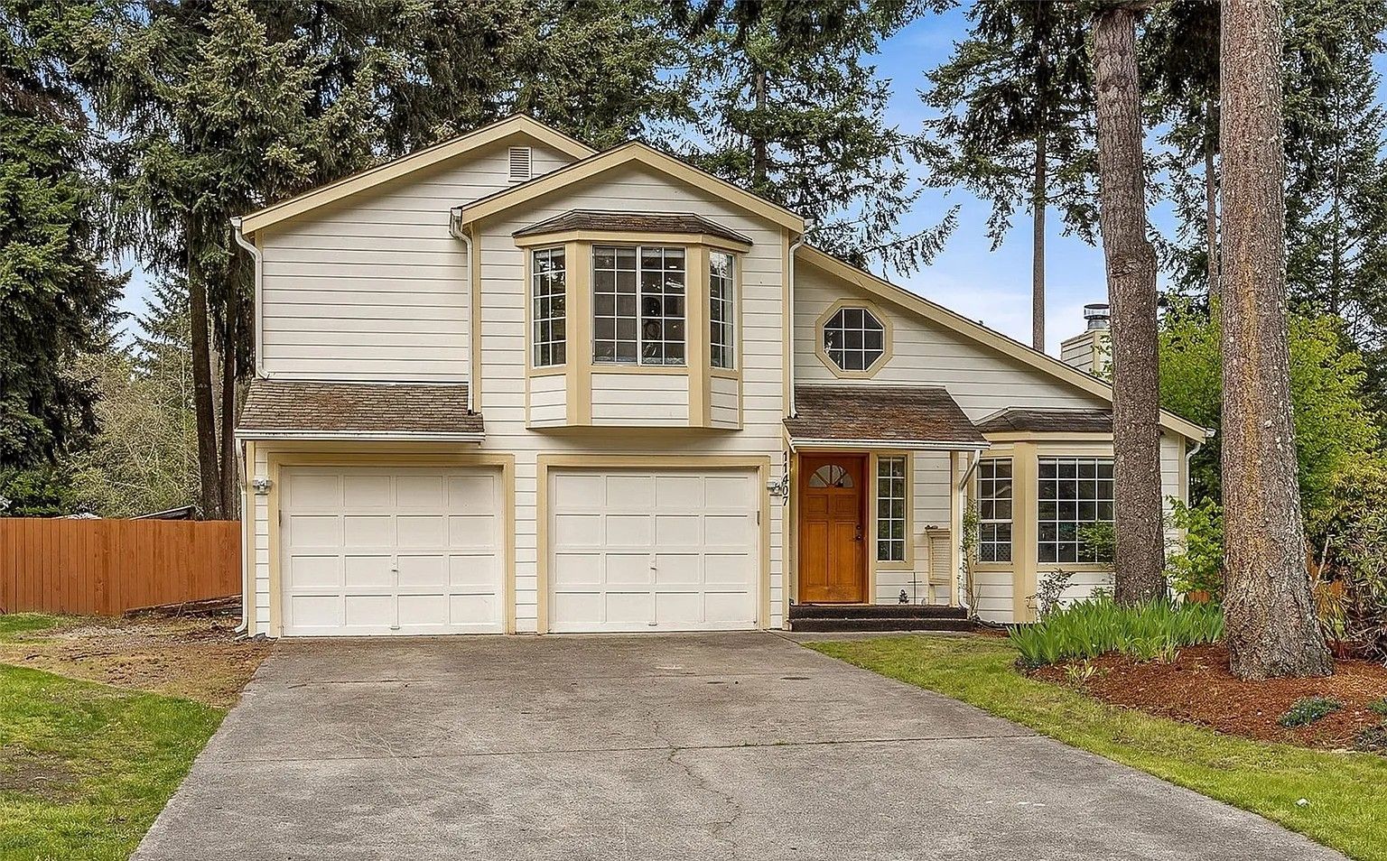 Suburban two-story house with beige siding, double garage, and a gravel driveway lined by tall trees.