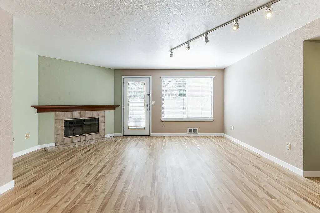 Empty living room with wood flooring, fireplace, door, and window. Light walls and ceiling.