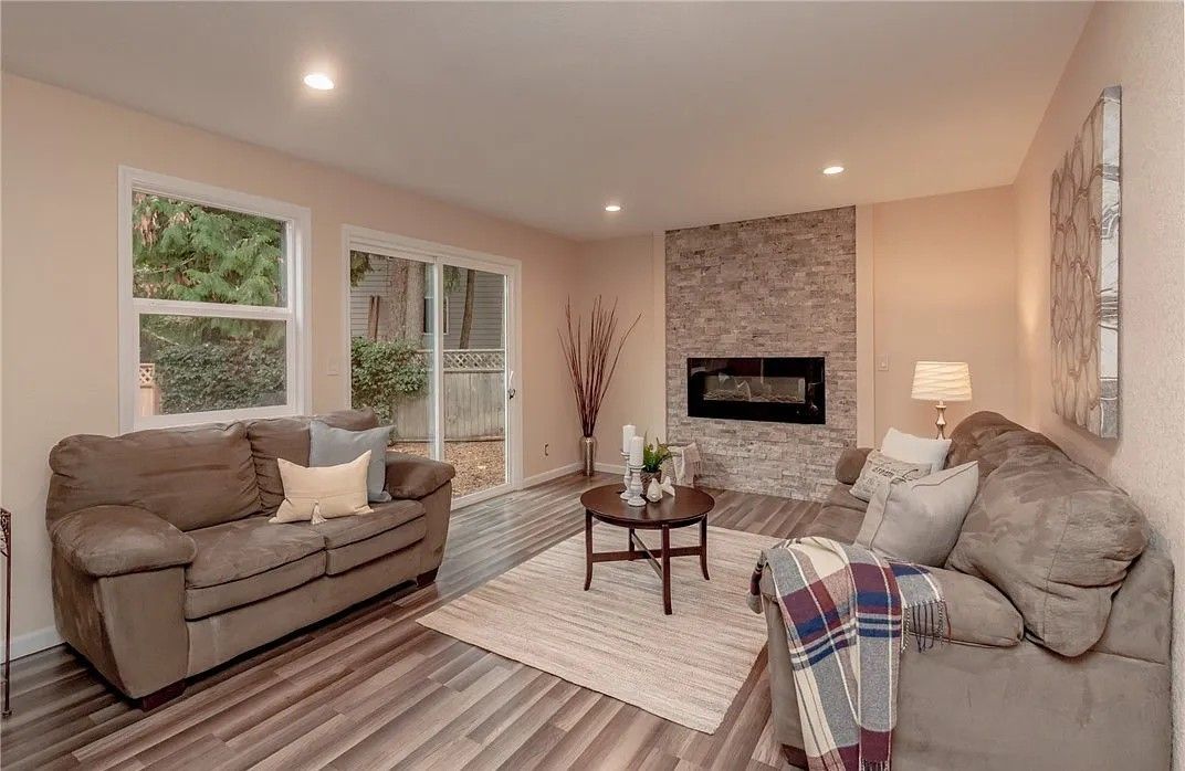 Cozy living room with fireplace, two sofas, a rug, and a small table. Two windows provide natural light.