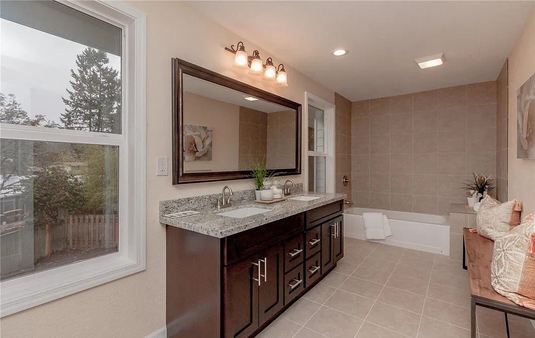 Bathroom with double sink vanity, large mirror, tub, and window overlooking trees. Brown cabinets and neutral tile.
