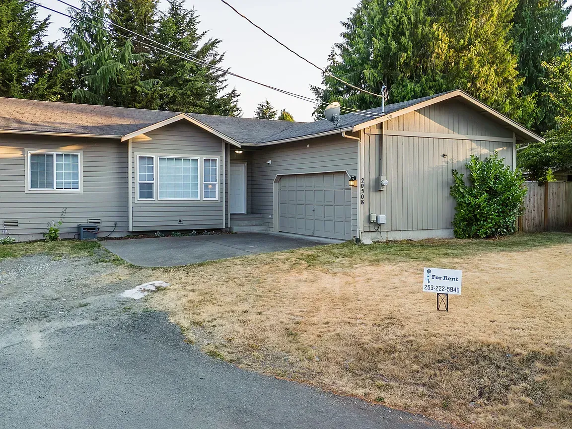 Gray house with garage, brown roof, dry lawn, trees in the background. A for sale sign in yard.