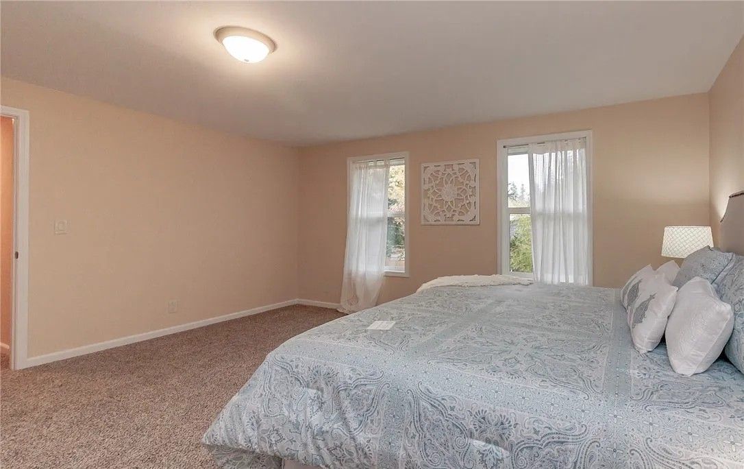 Bedroom with a large bed, two windows with sheer curtains, beige walls, and brown carpet.