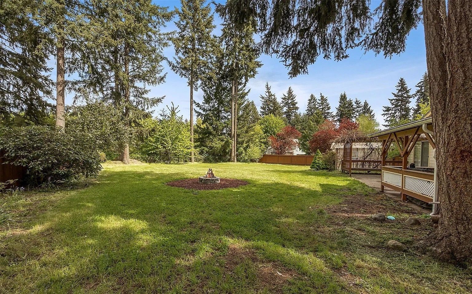 Backyard lawn with tall trees, a fire pit in the center, and a wooden bench on the right.