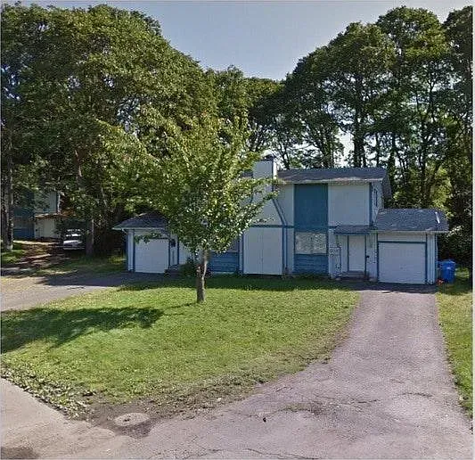 Two-story house with blue trim, white walls, and a driveway, surrounded by trees and a green lawn.
