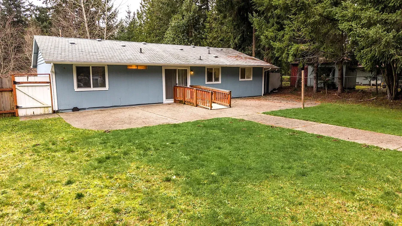 Blue house with a gravel patio and wooden ramp, surrounded by green grass and trees.