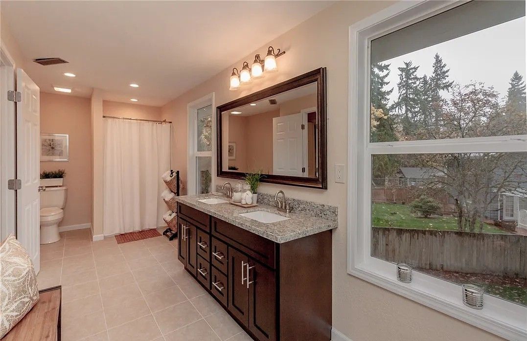 Bathroom with a double vanity, large mirror, and a window overlooking greenery. Beige walls and tiled floor.